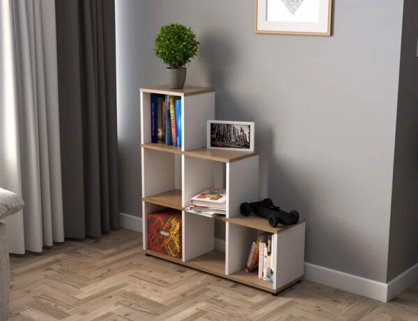 A small, tiered Multipurpose Stepping Bookcase with six cubby holes holds books, a photo frame, a plant pot with a small tree, and a pair of dumbbells. Behind the bookcase is a gray wall adorned with framed artwork, and a partially visible curtain drapes to the left.