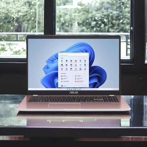 A sleek 15” Asus Intel Platinum 4GB/128 SSD laptop with a Windows 11 home screen displayed is placed on a glossy table. The background shows a window with a blurred view of greenery outside. The laptop's screen shows various app icons on a vibrant blue abstract background.